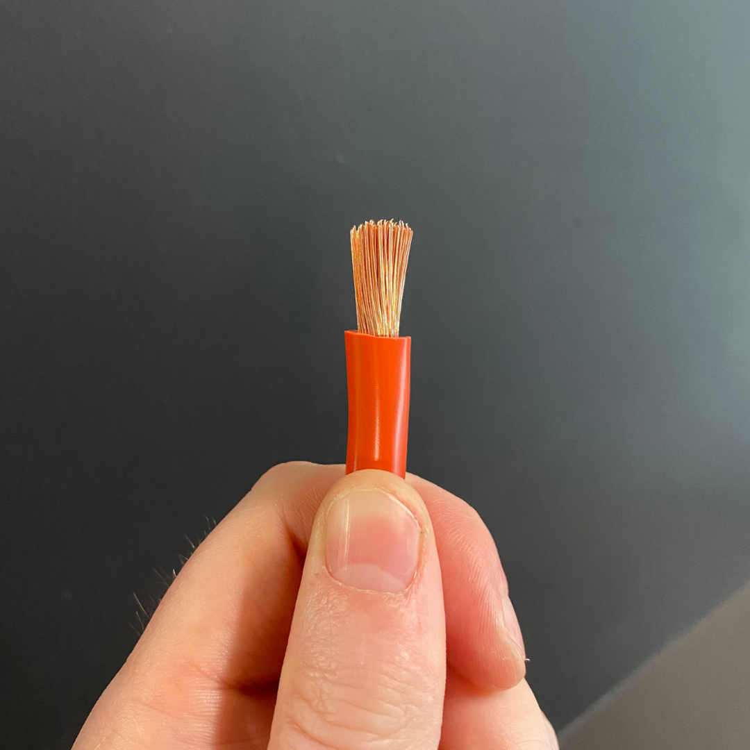 A close-up of a hand holding a 255mm battery cable electrical wire or cable connector with a bright orange outer jacket and multiple fine copper-colored strands exposed at the tip, against a dark gray background.