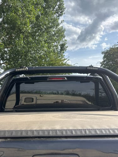 A black metal truck bed roll bar installed over the rear window of a pickup truck, with a beige tonneau cover on the bed. The background shows green trees and a cloudy blue sky.